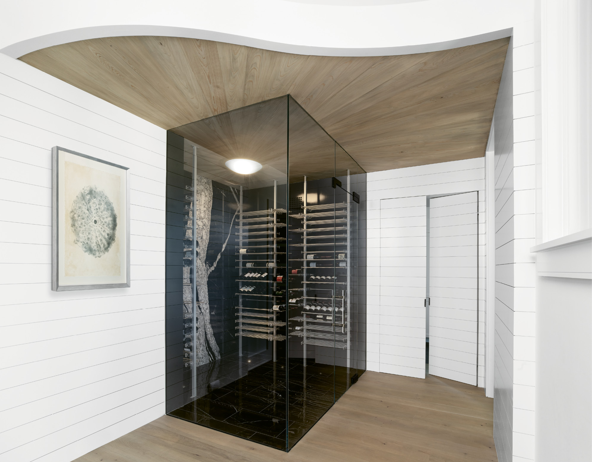The wine room features frameless glass doors, acrylic and anodized aluminum racks, and its own HVAC system. A granite slab covers the back wall for a dramatic statement, and a vintage light fixture anchors a corner from which cypress planks radiate.
